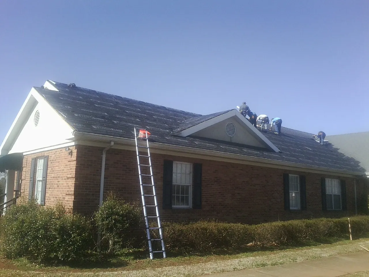 Crew tearing shingles off a brick ranch roof with a ladder set — Metal Roof Repair work by our crew in Montezuma